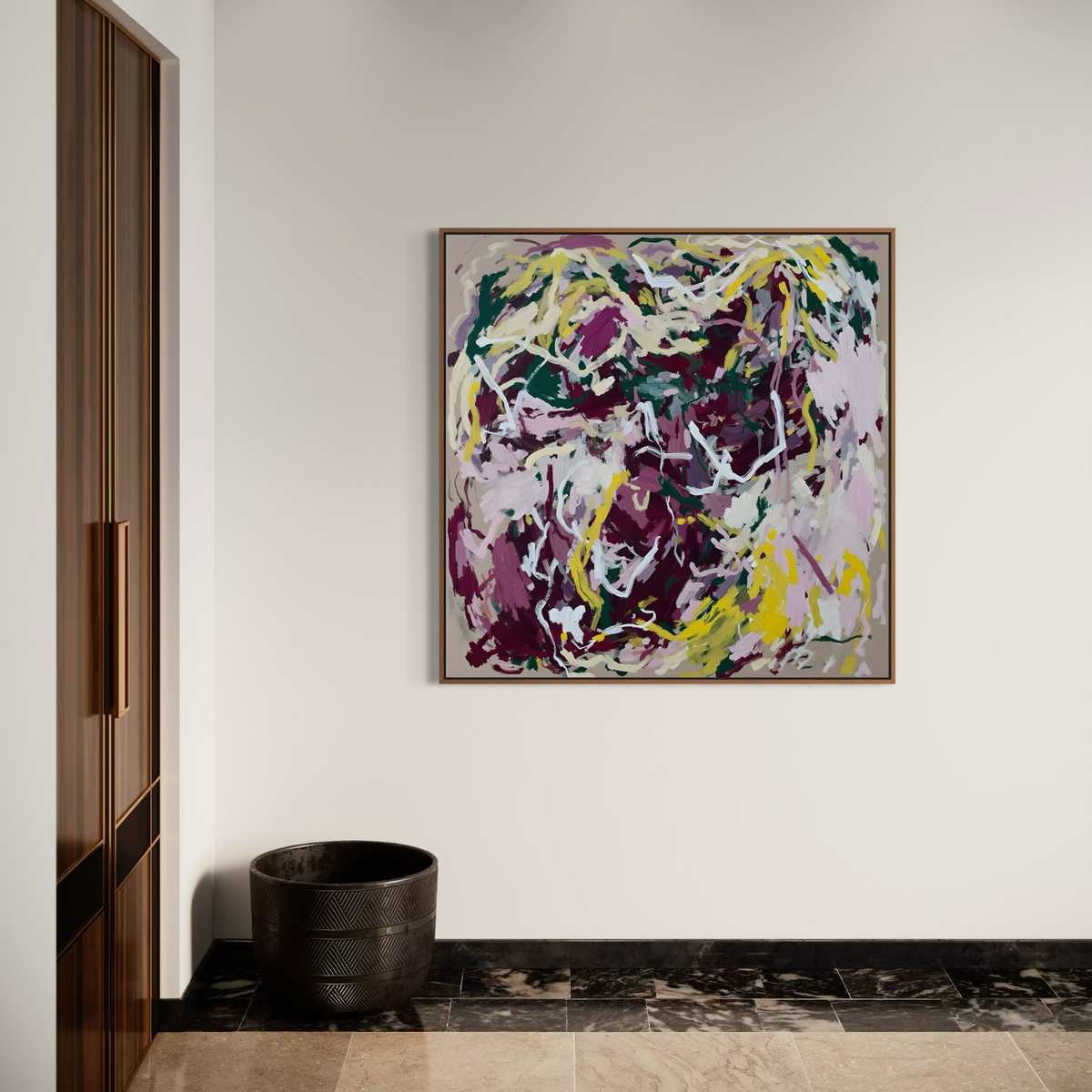 A square framed abstract painting with expressive brushstrokes in purple, yellow, green, white, and pink, hanging on a white wall in a modern interior setting with a dark wooden cabinet and a black textured planter.